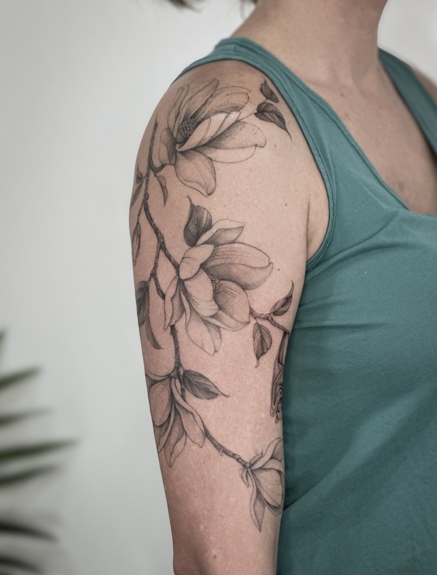 A delicate black and grey magnolia flower tattoo on the upper arm with detailed floral design — by Melanie Benoit, los angeles