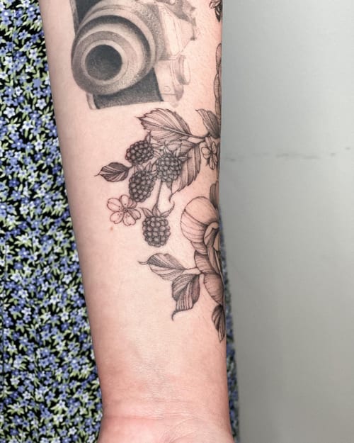 A delicate black and grey tattoo of a camera with floral and berry elements on the forearm, done in an illustrative style — by Melanie Benoit, los angeles