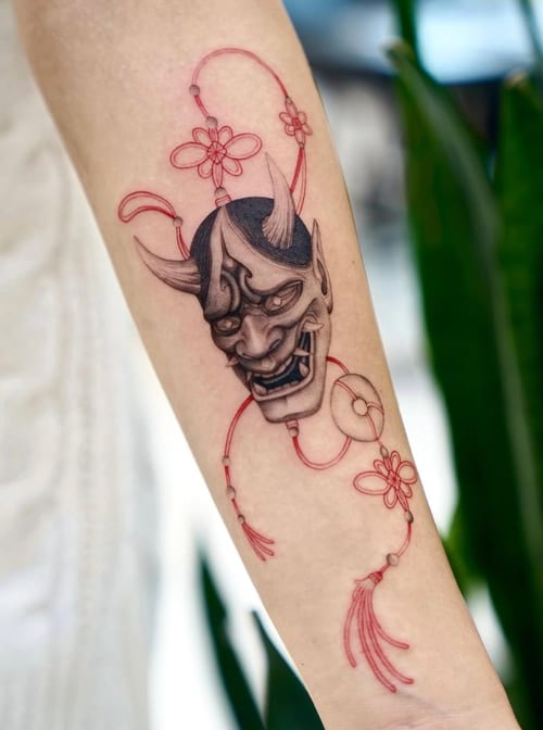 A fine line Japanese style tattoo of a demon mask on the forearm, featuring red and black ink with delicate floral elements — by otaybeatdown, los angeles