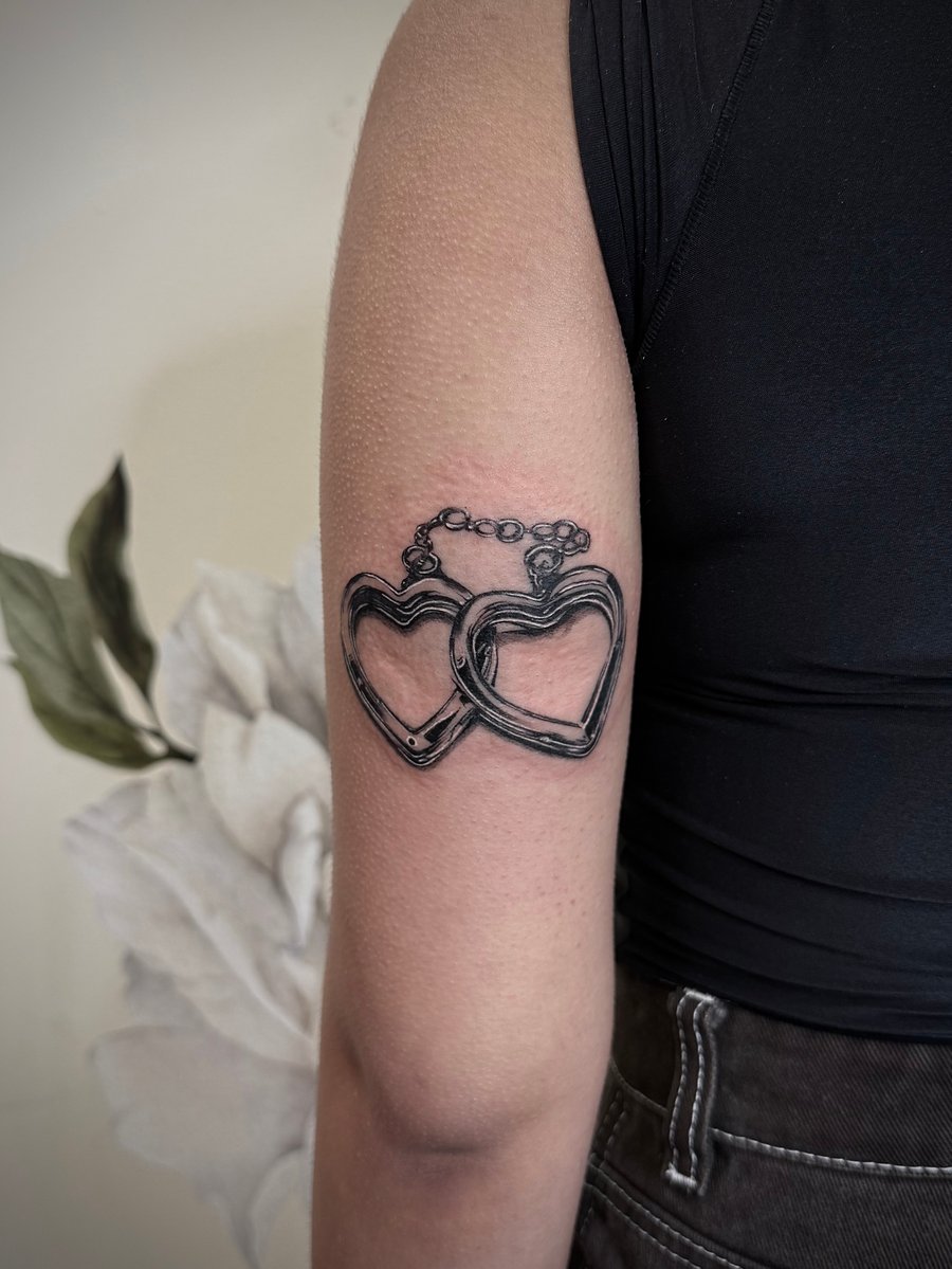 A black and grey tattoo of two interlocking heart-shaped chains on the upper arm, featuring bold linework and shading — by emily, los angeles