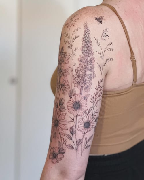A delicate black and grey floral arrangement with a bee on the upper arm, featuring fine line botanical details — by Melanie Benoit, los angeles