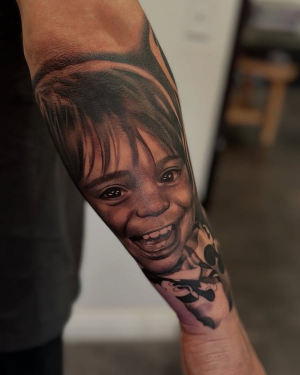 A detailed black and grey portrait of a child's face on the forearm, capturing a joyful expression with realistic shading — by Johnny Ayala, fontana, ca