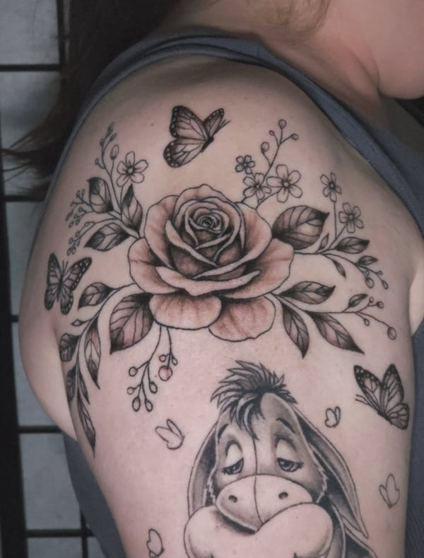 A black and grey tattoo featuring a rose, butterflies, and Eeyore on the shoulder, with delicate illustrative details — by Miguel ANGELO, temecula, ca