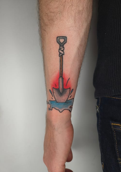 A colorful new school tattoo of a shovel on the forearm, featuring bold outlines and a splash of red and blue background — by ashynucklestattoo, los angeles