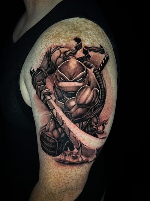 A black and grey realism tattoo of a Ninja Turtle wielding a sword on the upper arm, featuring detailed shading and dynamic pose — by Travis Ross, los angeles