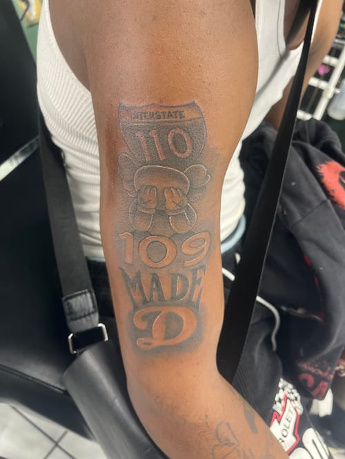 A black and grey tattoo on the upper arm featuring bold lettering and a shield-like design with the number 110 — by Mr._Folks, los angeles