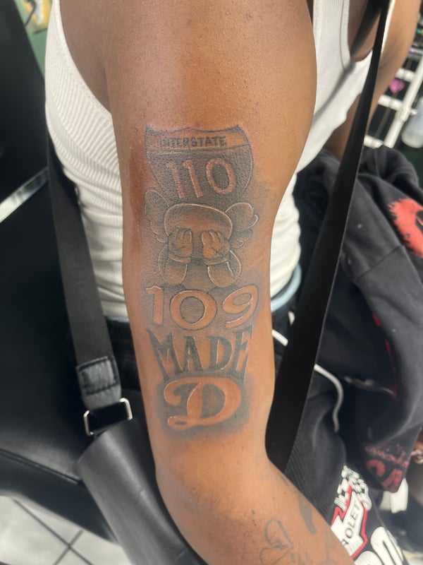 A black and grey tattoo on the upper arm featuring bold lettering and a shield-like design with the number 110 — by Mr._Folks, los angeles