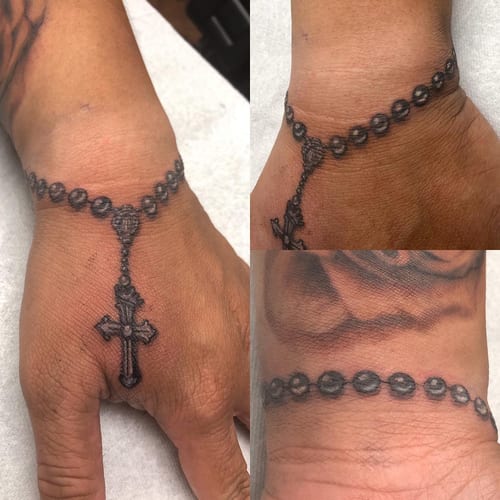 A black and grey rosary bracelet tattoo on the hand, featuring a detailed cross and beads in a realistic style — by Sandra Price, los angeles