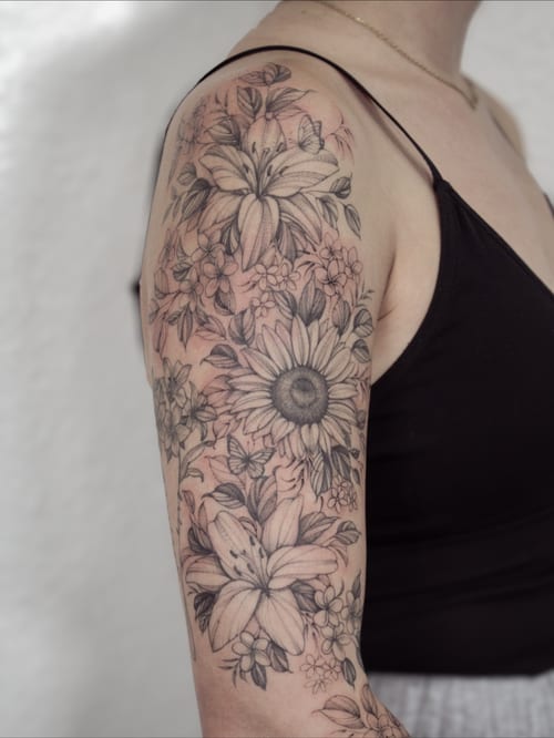 A delicate black and grey floral tattoo on the upper arm featuring lilies, sunflowers, and butterflies — by Melanie Benoit, los angeles