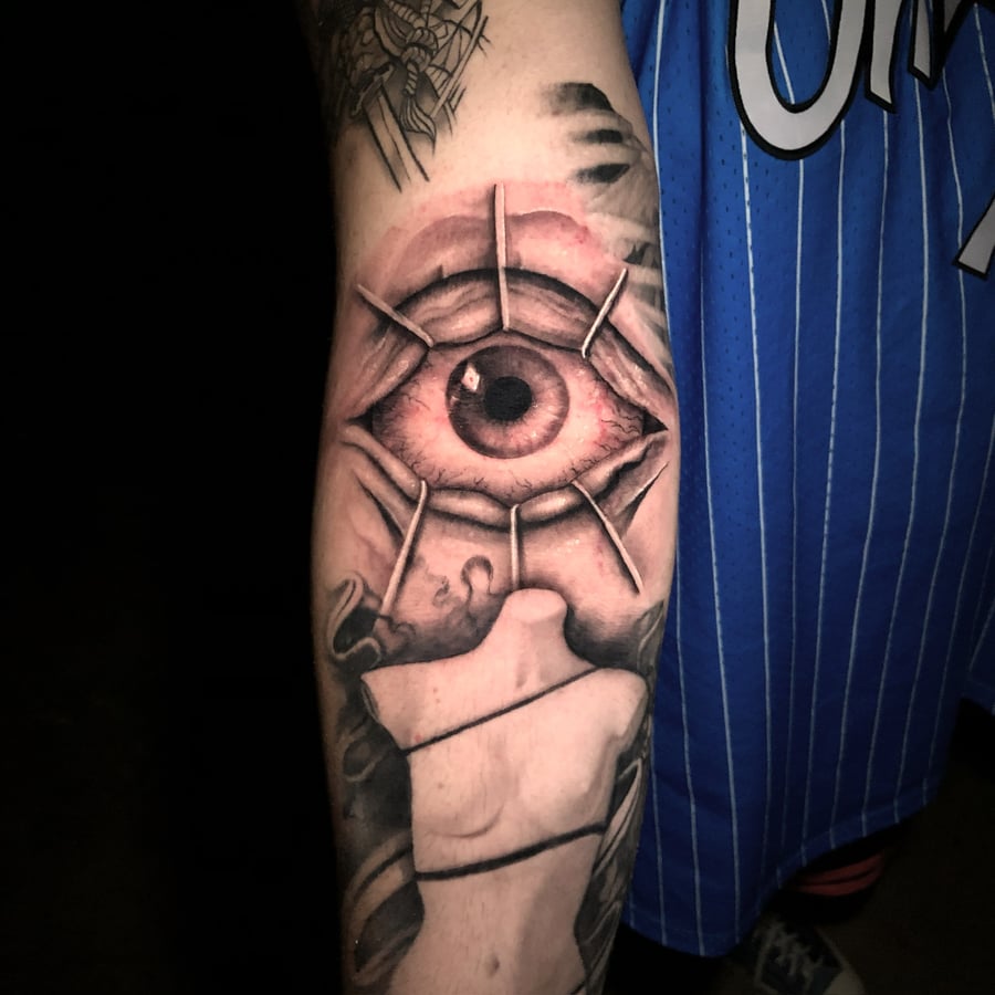A realistic tattoo of an eye on the forearm, featuring detailed shading and a mix of color and black ink — by Travis Ross, los angeles