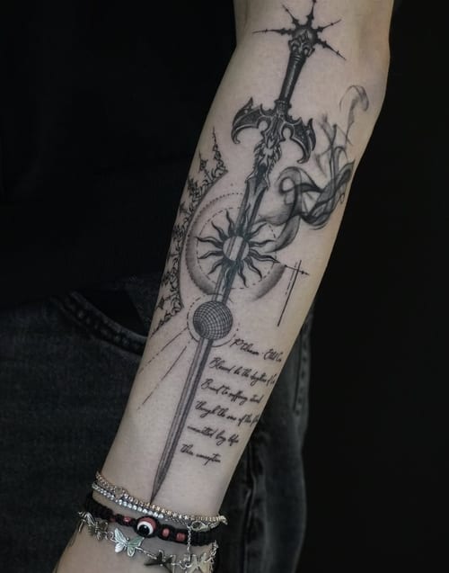 A black and grey illustrative tattoo on the forearm featuring an ornate sword with intricate linework and accompanying script — by Jian Liang, los angeles