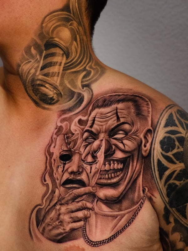 A black and grey tattoo of a man holding a cracked mask on the shoulder, featuring realistic details and a dark, graphic style — by Johnny Ayala, fontana, ca