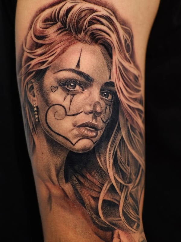A detailed black and grey woman's portrait with clown makeup on the upper arm, featuring realistic shading and Chicano style elements — by Johnny Ayala, fontana, ca
