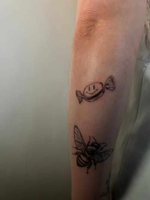 A minimalist black and grey tattoo on the forearm featuring a bee and a candy with a smiley face — by Brian Kies, los angeles