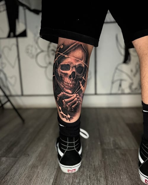 A realistic black and grey tattoo of a hooded skeleton on the calf, featuring detailed shading and a dark, graphic style — by zealous, los angeles