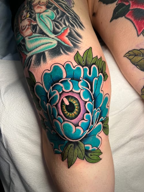A colorful Neo-Traditional tattoo of an eye surrounded by floral elements on the knee, featuring bold outlines and vibrant colors — by Megan Rose, los angeles