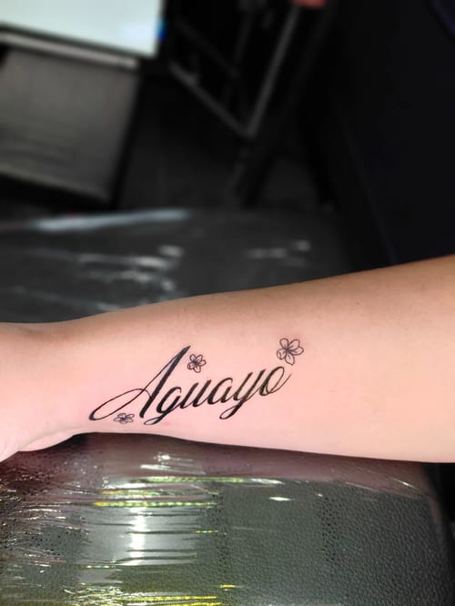 A black and grey lettering tattoo on the forearm featuring the word 'Aguayo' with small floral accents — by anoetic.designs.ultd, los angeles