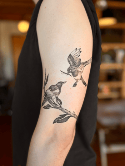 A black and grey fine line tattoo of two birds on a branch on the upper arm, featuring realistic details and soft shading — by Dylan Aiello, los angeles