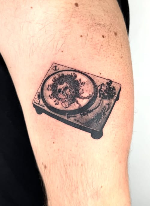 A black and grey illustrative tattoo of a turntable with a skull in the center, located on the upper arm — by Level tattoo, los angeles