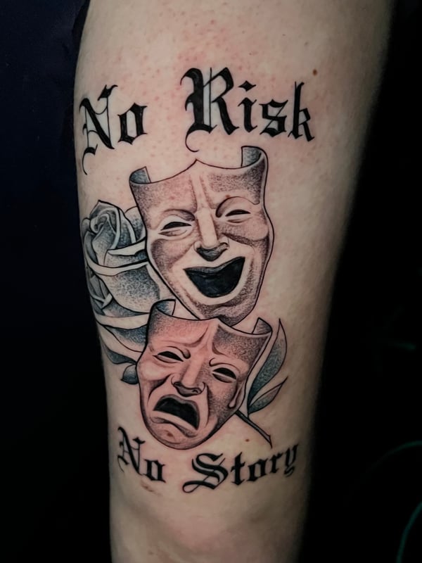 A black and grey tattoo on the arm featuring theatrical masks with a rose and script reading 'No Risk No Story' — by Josue Bravo, los angeles