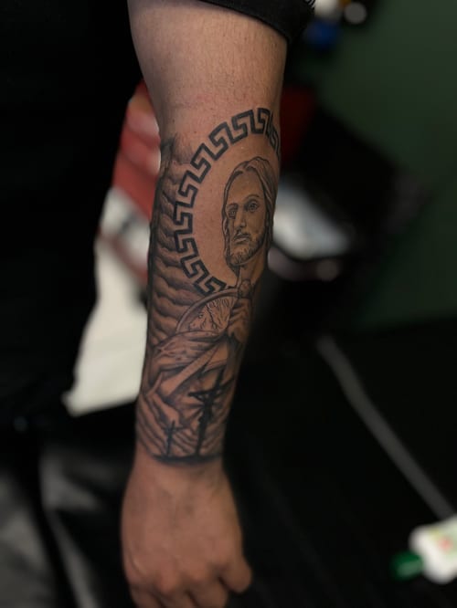 A black and grey realism portrait tattoo on the forearm featuring a male figure with detailed shading and a Greek key pattern border — by Mr._Folks, los angeles