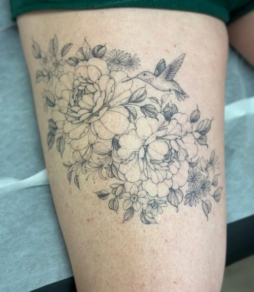 A delicate black and grey tattoo of a hummingbird with a floral arrangement on the thigh, featuring fine line work and intricate details — by Melanie Benoit, los angeles