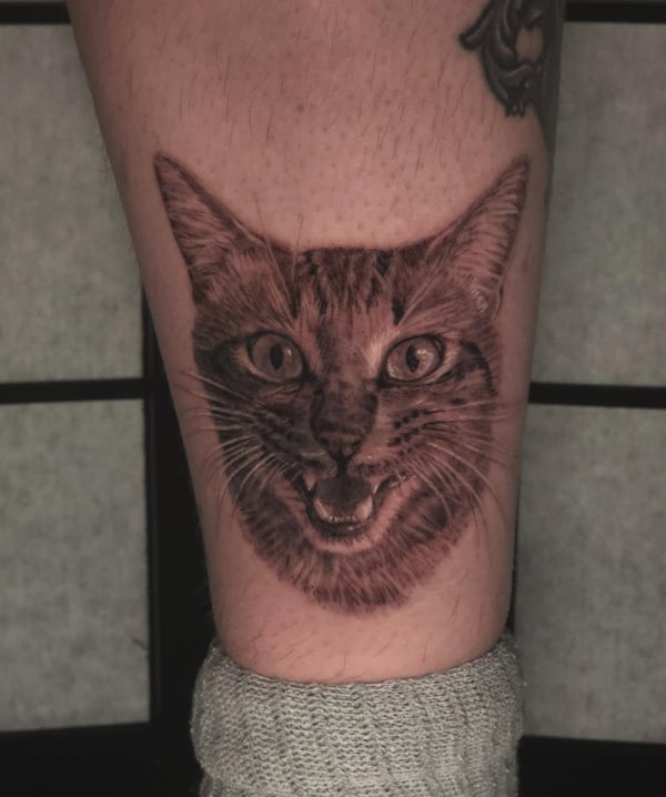 A realistic black and grey cat portrait tattoo on the leg, capturing detailed fur and expression — by Miguel ANGELO, temecula, ca