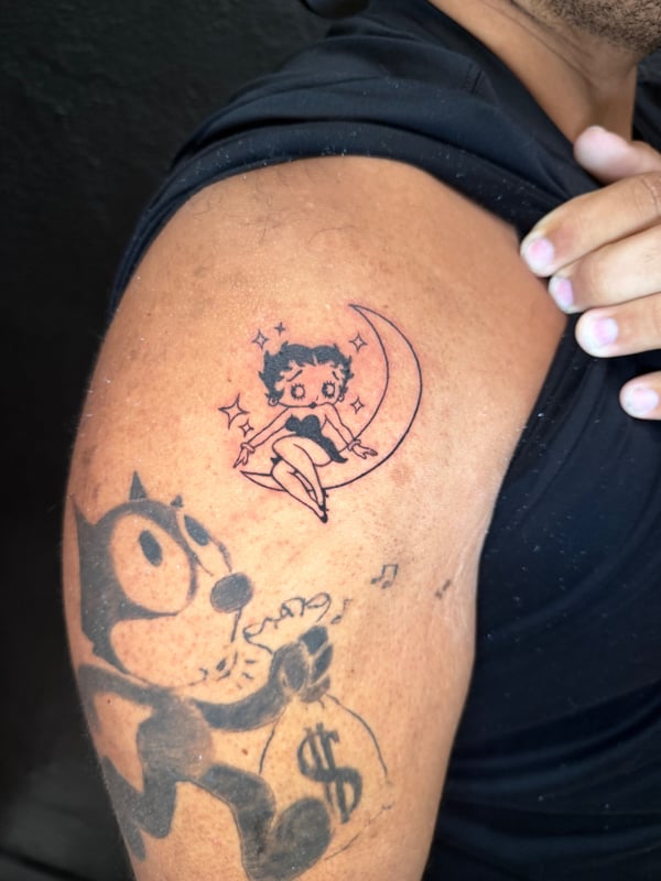 Cartoon characters tattooed in black linework on the upper arm, featuring a character sitting on a crescent moon with stars — by Kiera Garvin, los angeles