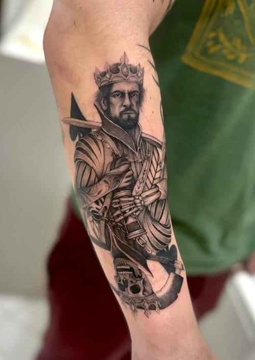 A detailed black and grey realism tattoo of a crowned man in armor on the forearm, featuring intricate shading and texture — by otaybeatdown, los angeles