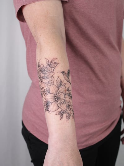 A delicate black and grey floral tattoo with a hummingbird on the forearm, featuring fine line work and clean details — by Melanie Benoit, los angeles