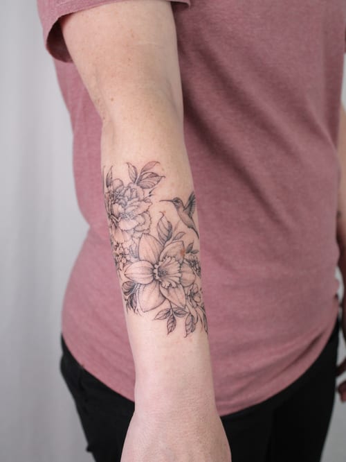 A delicate black and grey floral tattoo with a hummingbird on the forearm, featuring fine line work and clean details — by Melanie Benoit, los angeles