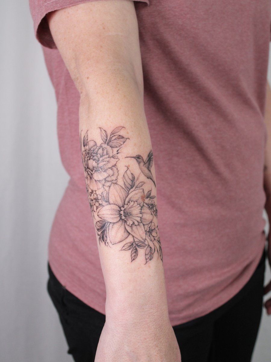 A delicate black and grey floral tattoo with a hummingbird on the forearm, featuring fine line work and clean details — by Melanie Benoit, los angeles