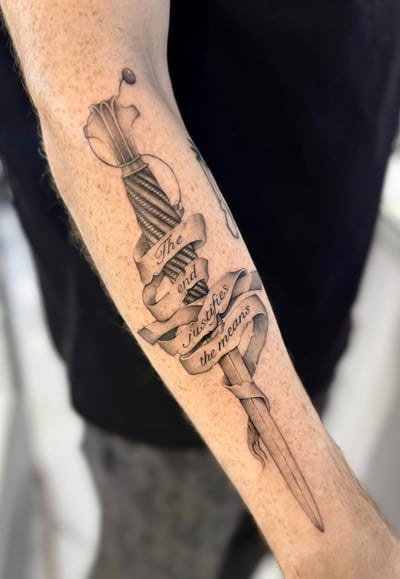 A black and grey linework tattoo of a dagger wrapped with a ribbon featuring script on the forearm — by otaybeatdown, los angeles