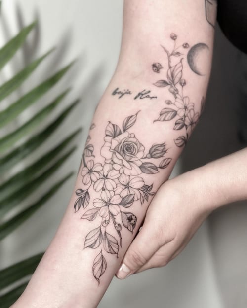 A delicate black and grey floral arrangement with roses and script on the forearm, featuring fine line work and minimalistic design — by Melanie Benoit, los angeles