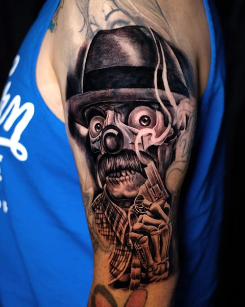 A black and grey Chicano-style tattoo of a clown with a gun and skeleton hand on the upper arm — by Johnny Ayala, los angeles