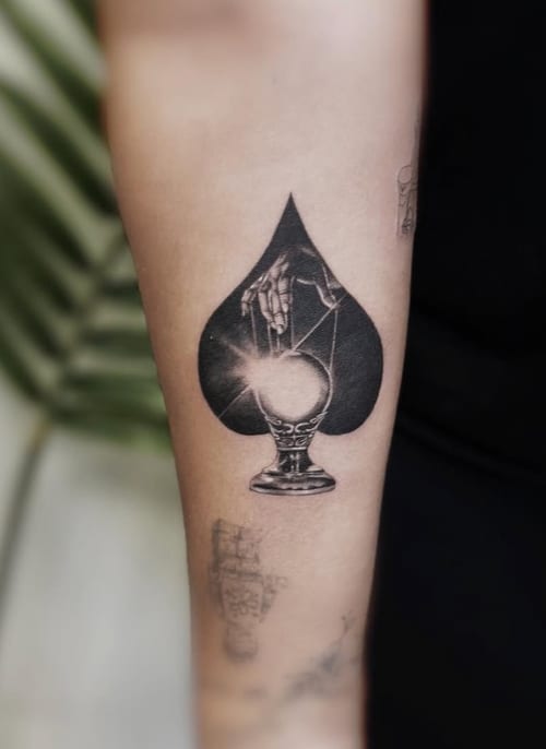 A black and grey surrealistic tattoo on the forearm featuring a skeletal hand holding a crystal ball within a spade shape — by Jian Liang, los angeles