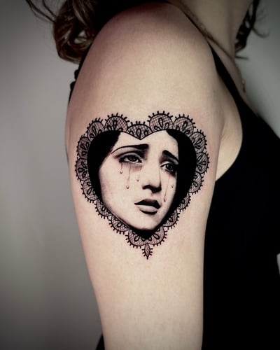 A black and grey portrait of a woman's face with tears, framed by an ornamental lace heart on the upper arm — by emily, los angeles