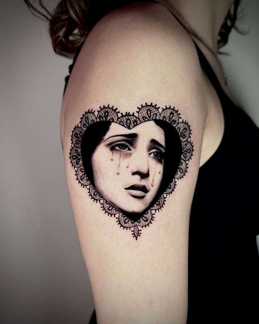 A black and grey portrait of a woman's face with tears, framed by an ornamental lace heart on the upper arm — by emily, los angeles