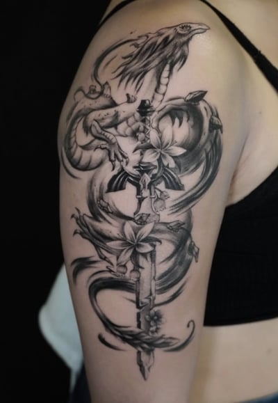 A black and grey illustrative tattoo on the upper arm featuring a dragon intertwined with flowers and a sword, showcasing detailed linework and shading — by Jian Liang, los angeles