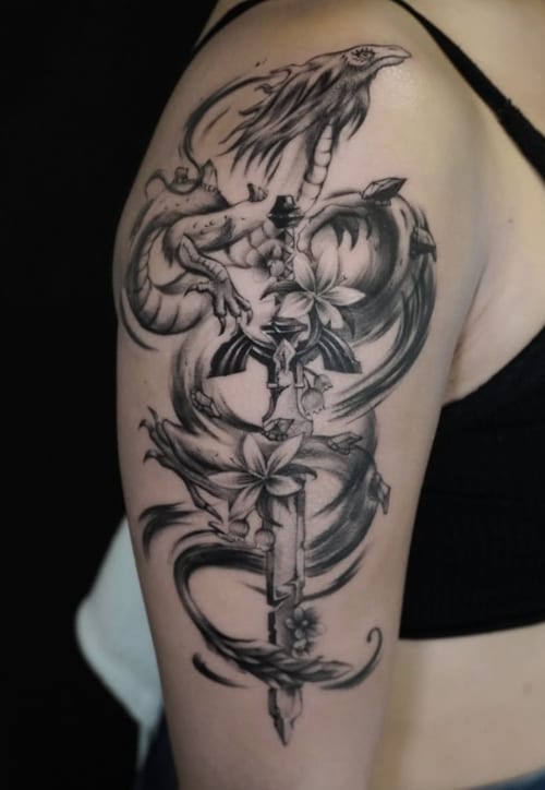 A black and grey illustrative tattoo on the upper arm featuring a dragon intertwined with flowers and a sword, showcasing detailed linework and shading — by Jian Liang, los angeles