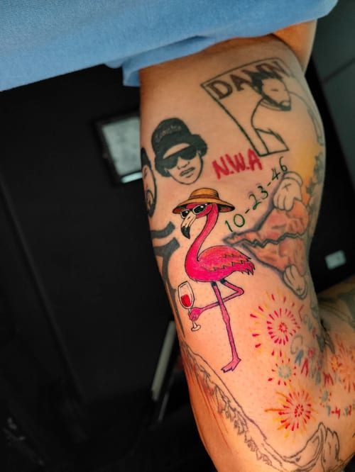 A colorful illustrative tattoo of a flamingo wearing sunglasses and a hat, holding a wine glass, with script and additional graphic elements on the arm — by anoetic.designs.ultd, los angeles