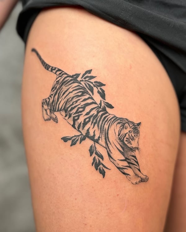 A bold blackwork tattoo of a tiger on the thigh, featuring an illustrative style with leaves — by Dylan Aiello, los angeles