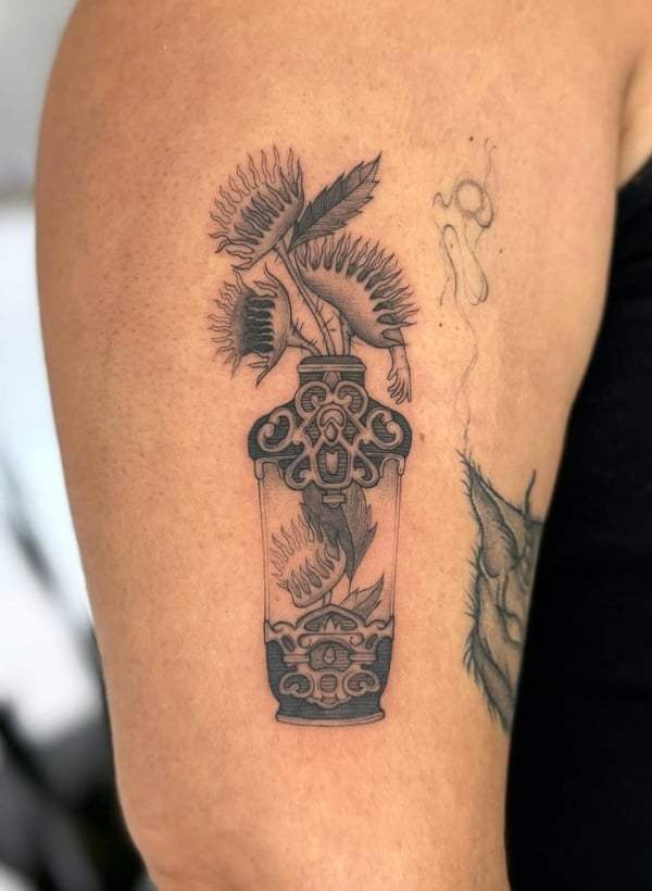 A black and grey ornamental tattoo of a decorative vase with plants on the upper arm, featuring intricate patterns and shading — by otaybeatdown, orange, ca