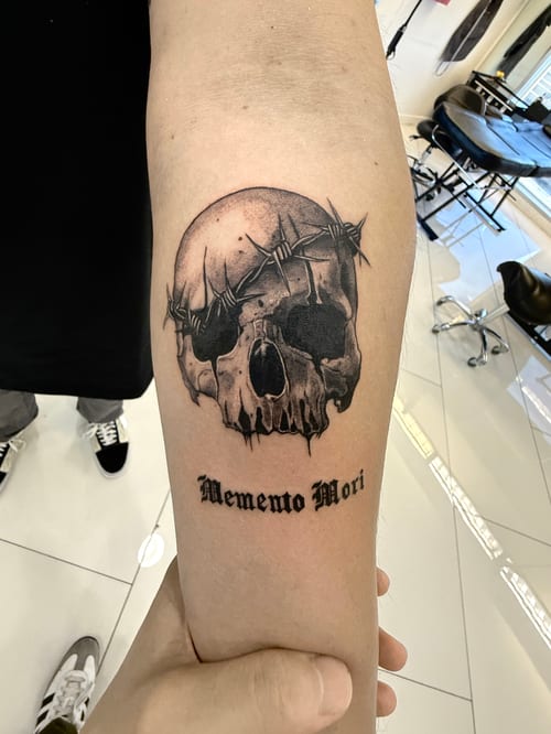A black and grey realism tattoo of a skull wrapped in barbed wire with the script 'Memento Mori' on the forearm — by 钟逸政, los angeles
