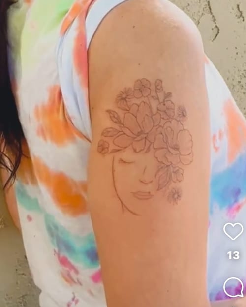 A delicate black and grey tattoo of a woman's face with flowers on the upper arm, featuring fine line floral details — by Melanie Benoit, los angeles