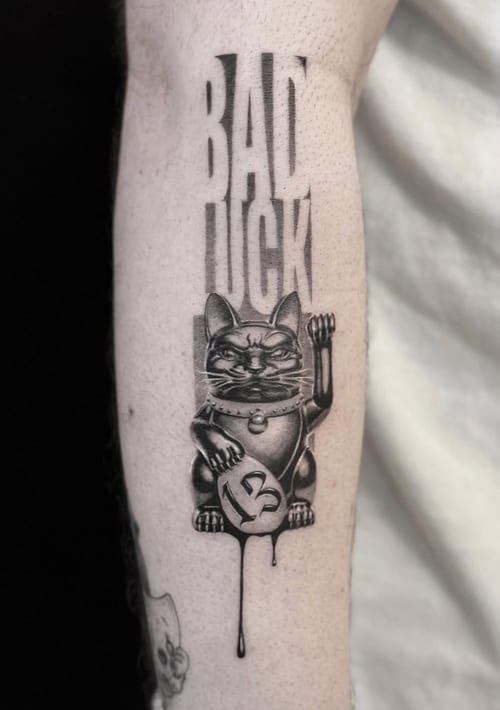 A black and grey illustrative tattoo of a lucky cat with the number 13 and the words 'Bad Luck' on the forearm, featuring bold lines and shading — by Jian Liang, los angeles