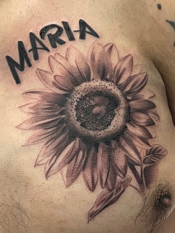 A realistic black and grey sunflower tattoo on the chest with the name 'Maria' in bold script above it — by Josue Bravo, los angeles