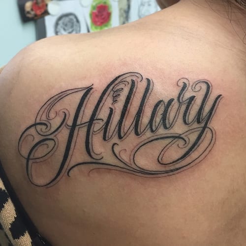 A bold black and grey script lettering tattoo of the name 'Hillary' on the shoulder — by Johnny Ayala, los angeles
