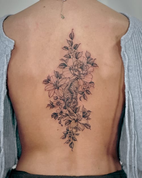 A delicate black and grey fine line tattoo of a tiger surrounded by flowers on the back — by Melanie Benoit, los angeles