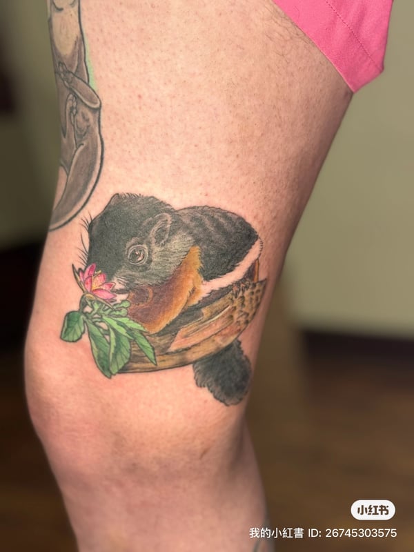 A realistic color tattoo of a squirrel holding a flower on the leg, featuring detailed fur and vibrant colors — by Sandra Price, ventura, ca